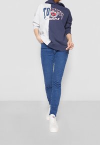 Felpa con cappuccio blu e grigia con un grande logo "TOMMY", abbinata a jeans blu e sneakers bianche. La felpa presenta un design a contrasto.
