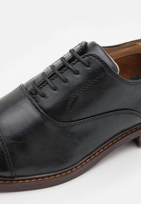 Chaussure en cuir noir avec une finition lisse, dotée d'un bout arrondi, d'un laçage et de coutures contrastantes sur la semelle marron.