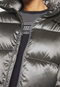 Grå puffjacka med glänsande yta, med huva och dragkedjestängning, lager på lager över en svart texturerad topp med logomönster.