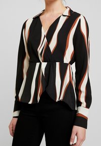 Blusa a righe bianco e nere con dettagli marroni, caratterizzata da un design avvolgente, maniche lunghe e un dettaglio in vita a peplo.