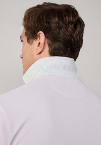 Světle fialová polo košile se texturovaným světle modrým límcem, na kterém je nápis "HACKETT" v růžové barvě. Viditelné švy a hladká textilie.