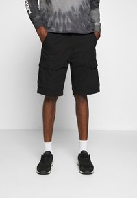 Shorts cargo noirs avec poches latérales, longueur au-dessus du genou, associés à des chaussettes blanches et des chaussures de sport noires. Modèle portant un haut gris tie-dye.