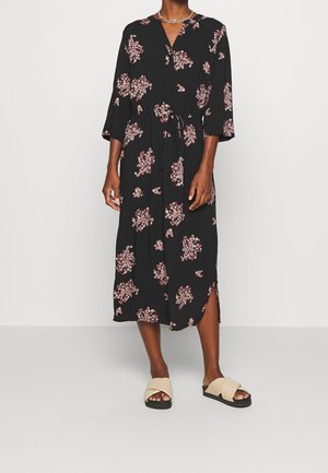 Femme portant une robe midi noire avec des motifs floraux roses, des manches trois-quarts, une ceinture à la taille et des sandales beiges sans lacets.