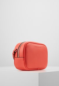 Korallfärgad faux läder crossbody-väska, rektangulär form, toppdragkedja, sidorems, präglad logotyp, slät textur med rundade kanter.