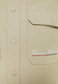 Tessuto di corduroy beige con bottoni a pressione e una tasca a patta ricamata con "Tommy Hilfiger EST. 1985" in filo bianco, rosso e blu navy.