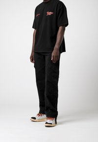 Zwarte katoenen t-shirt met rode grafische tekst en een relaxte fit, gecombineerd met zwarte cargobroek met diagonale naden en witte sneakers met kleurrijke veters.