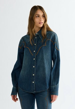 Frau trägt dunkelblaue Denim-Hemd und passende Jeans mit dekorativer Naht, steht und schaut seitlich vor einfachem Hintergrund.