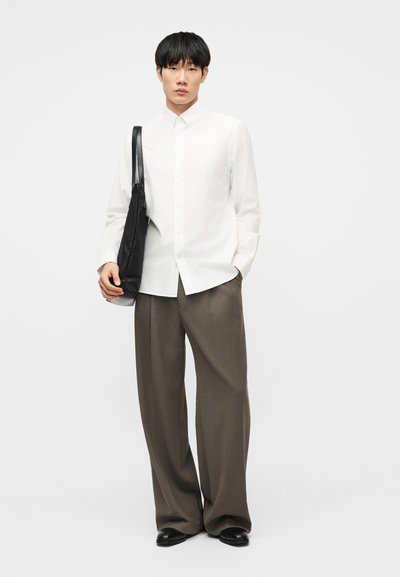 Άνδρας με λευκό πουκάμισο με κουμπιά, φαρδιές καφέ παντελόνες, μαύρα παπούτσια και μαύρη τσάντα ώμου, στέκεται μπροστά σε απλό λευκό φόντο.