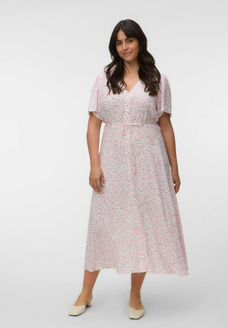 Femme aux longs cheveux foncés portant une robe midi blanche à imprimé floral rose avec ceinture nouée, debout devant un fond uni.