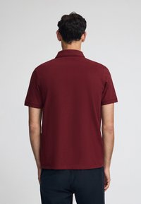 Burgundy polo skjorta med klassisk krage, korta ärmar och en mjuk textur. Baksidan har en ren design utan utsmyckningar.