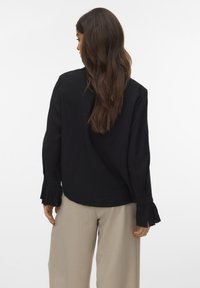 Black textured blouse with long sleeves and ruffled cuffs, worn with beige high-waisted pants, viewed from the back.