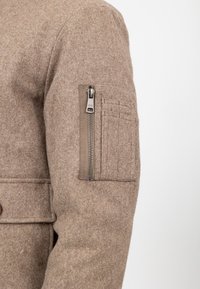 Braune Wolljacke mit einem vertikalen Reißverschlussfach und einer gestickten rechteckigen aufgesetzten Tasche am Oberarm vor einem weißen Hintergrund.