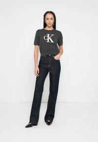 Tricou gri cu mâneci scurte și logo-ul "CK", asortat cu blugi înalți, de culoare albastru închis. Modelul stă cu o mână în buzunar, purtând balerini negri cu vârf ascuțit.