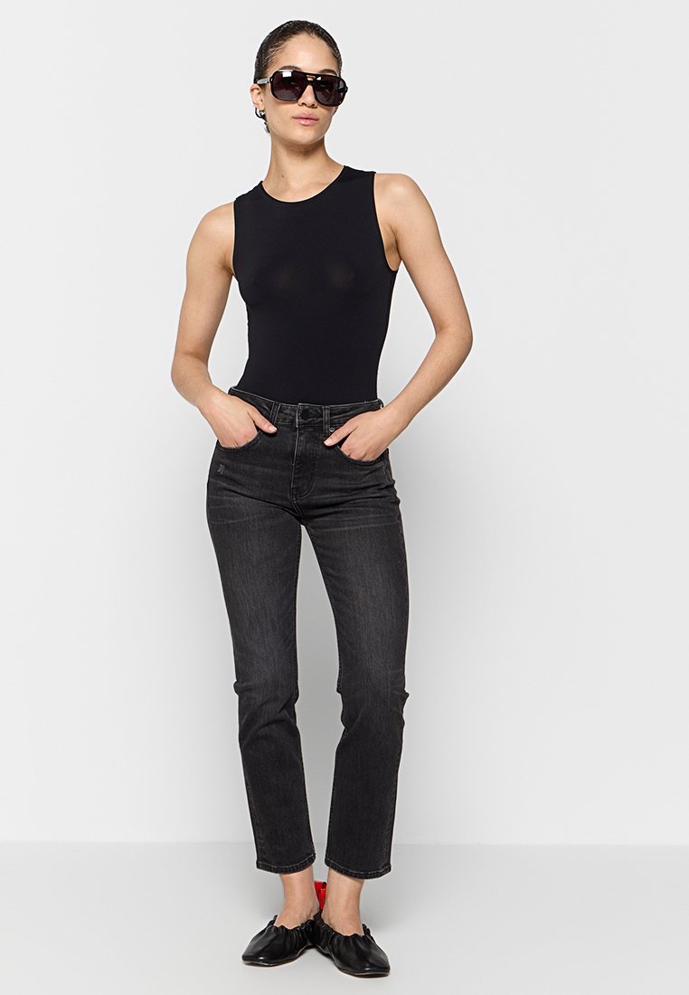 Kobieta w dużych czarnych okularach przeciwsłonecznych, bezrękawnik czarny, ciemne jeansy o wysokim stanie oraz czarne baleriny z czerwonymi akcentami na obcasie.