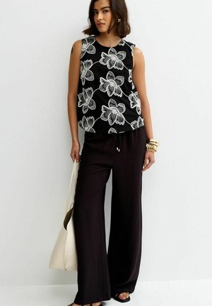 Donna con top nero senza maniche con grande stampa floreale bianca, pantaloni neri a gamba larga, che tiene una borsa tote beige e indossa braccialetti e orecchini dorati.