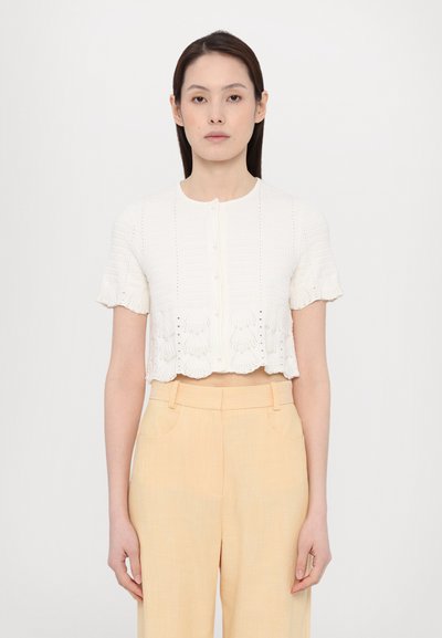 Weißes cropped Blusenshirt mit kurzen Ärmeln, strukturiertem Muster, Knopfleiste und gewelltem Saum. Kombiniert mit hellgelben, taillierten Hosen.