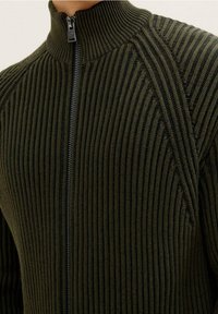 Stickad zip-up-tröja i mörkgrön, med hög krage, ribbad textur och kontrasterande vertikala ränder längs axlarna.