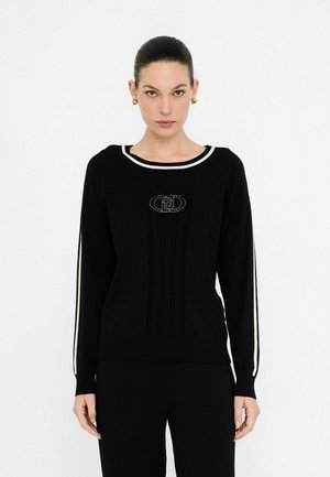 Donna che indossa un maglione nero a maniche lunghe con strisce bianche sulle maniche e sul colletto, con un piccolo logo di cristallo sul petto.