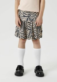 Krilo s črtastim vzorcem zebra v bež in črni barvi, izdelano iz mehkega blaga. Kombinirano z belimi nogavicami do kolen in črnimi lakastimi loaferji.