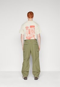 Beige t-shirt with red graphic print on the back, paired with loose olive green cargo pants featuring side pockets.