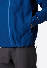 Osoba ve modré bundě se zipem vpředu a rukou v kapse, v kombinaci s tmavě šedými kalhotami, na bílém pozadí.