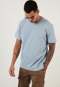 Tricou din bumbac de culoare albastru deschis, cu guler rotund și mâneci scurte, purtat cu pantaloni cargo maro. Afișând tatuaje complicate pe brațe.