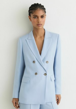 Kvinde med flettet hår iført lyseblå dobbeltradet blazer med knapper og matchende bukser, stående foran en hvid væg.