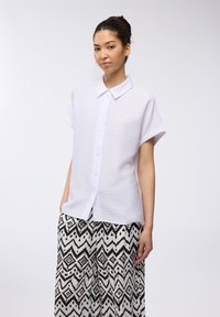 Camicetta bianca a maniche corte con finitura testurizzata, abbinata a pantaloni larghi a righe bianchi e neri con design geometrici.