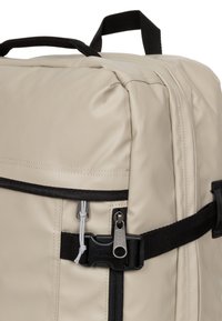 Beige ryggsäck tillverkad av slitstarkt material med svarta detaljer. Har en ficka med dragkedja, ett spännband och ett robust handtag på toppen.