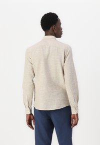 Camisa de manga larga beige claro en tejido texturizado, con cuello tipo solapa, puños abotonados y dobladillo curvo, combinada con pantalones azul oscuro.