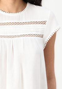 White blouse with short sleeves, featuring lace detailing and piped trim at the neckline and sleeve hems. Lightweight, textured fabric.