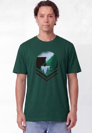 Homme portant un t-shirt vert foncé avec un graphisme d'un vététiste près des arbres et d'un lac dans un design circulaire à l'avant.