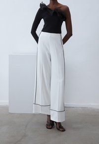 Top nero a spalla unica con dettagli a volant abbinato a pantaloni bianchi a gamba larga a vita alta con piping nero. Sandali neri con tacco.