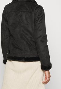 Zwarte nepbontjas met een gestructureerde afwerking, een gevoerde kraag en een ceintuur in de taille. Zachte fleece voering bij de manchetten.