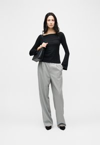 Γυναίκα που φορά μαύρο μπλουζάκι με μακριά μανίκια και ανοιχτό γκρι παντελόνι με ρίγες, κρατώντας μαύρη τσάντα ώμου, στέκεται σε ουδέτερο φόντο.