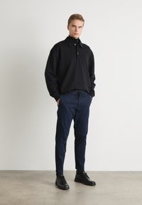 Black collared pullover, long sleeves, paired with navy drawstring trousers. Model wears black lace-up shoes on a neutral background.