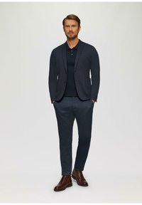 Blazer marine sur un polo noir, associé à un pantalon au motif longueur cheville. Des bottes en cuir marron à lacets complètent la tenue. Design simple et ajusté.
