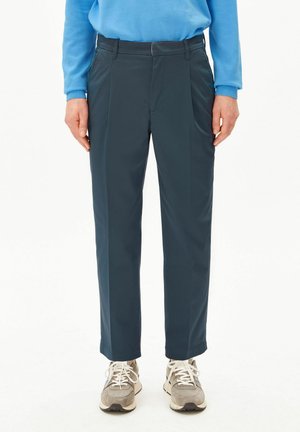 Personne portant un pantalon droit bleu marine, un sweatshirt bleu clair et des baskets grises à lacets, debout les bras détendus le long du corps.
