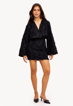 Femme portant une mini-robe noire texturée à manches longues avec un décolleté plongeant en V et des mules à talons hauts noires à bout pointu ornées de clous argentés.