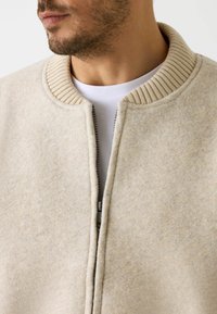 Fleece jack in lichtbeige met een geribde kraag, voorzien van een ritssluiting aan de voorkant en een zachte textuur, gestructureerd ontwerp zonder zichtbare patronen.
