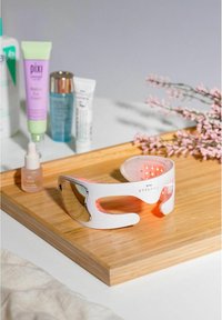 Masque facial LED blanc au design courbé et verres teintés, posé sur un plateau en bois aux côtés de produits de soin de la peau et de fleurs roses.