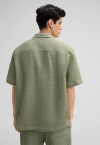Groen, kort-waardige shirt met een gestructureerd, wafelbreipatroon. Bevat een klassieke kraag en heeft een ontspannen pasvorm. Zichtbare rugnaaddetails.