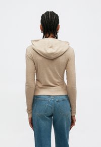 Μπεζ cropped φούτερ με άνετη εφαρμογή, διαθέτει κουκούλα, μακριά μανίκια και ριμπ τελείωμα. Συνδυάζεται με ψηλόμεσα μπλε τζιν.