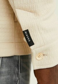 Poignet de chemise à manches longues texturée crème avec un bouton et une étiquette noire portant la mention « CHASIN », portée sur un jean denim foncé.