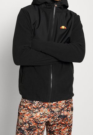 Osoba ubrana w czarną bluzę z kapturem na zamek błyskawiczny z pomarańczowym logo oraz kolorowe spodnie moro, ze skrzyżowanymi ramionami na jednolitym tle.