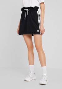 Žena v černé sukni Adidas s bílými pruhy, bílým šňůrkovým pasem, bílými ponožkami a bílými teniskami Adidas stojí na jednobarevném pozadí.