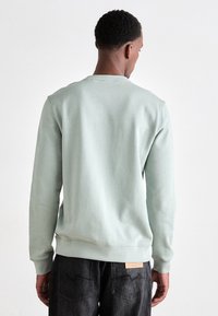 Man van achteren gezien, gekleed in een lichtgroene sweatshirt en zwarte jeans voor een effen witte achtergrond.