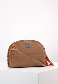 Hnědá kožená crossbody taška se šitými detaily, zaobleným tvarem, dlouhým popruhem a růžovými třásněmi. Stříbrná logová visačka na přední straně.