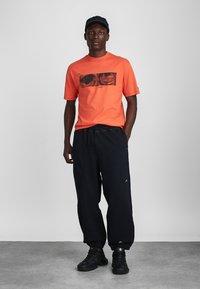 Πορτοκαλί βαμβακερό μπλουζάκι με γραφικό σχέδιο, συνδυασμένο με μαύρες φαρδιές φόρμες. Το μοντέλο φοράει ένα μαύρο καπέλο και μαύρα αθλητικά παπούτσια.