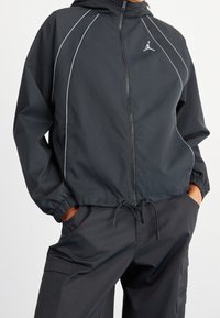 Svart sportjacka med dragkedja framtill, huva och elastiska ärmslut. Har ljusblå accenter och en liten logotyp på övre vänstra sidan.